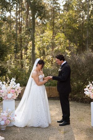Bride and groom exchange rings outdoors at a garden wedding ceremony surrounded by pink floral arrangements and tall trees.