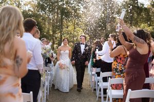 Bride and groom walk down outdoor aisle as guests toss confetti at a sunny woodland wedding ceremony.