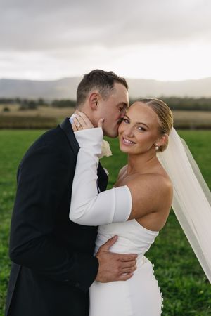Romantic outdoor wedding photo of newlyweds embracing in a sleek white dress and black suit against mountain landscapes.
