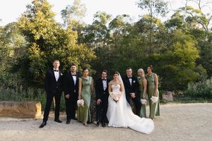 Wedding party in formal attire poses outdoors, groomsmen in tuxedos and bridesmaids in sage green gowns with bride and groom.
