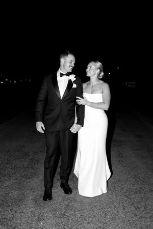 A black and white photo of a bride and groom walking together at night, sharing a laugh in formal wedding attire.