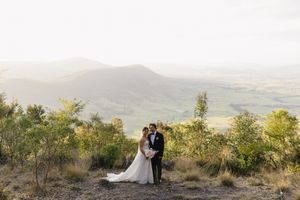Bride and groom embrace on rocky hilltop with sweeping green valley and misty mountain views behind them.