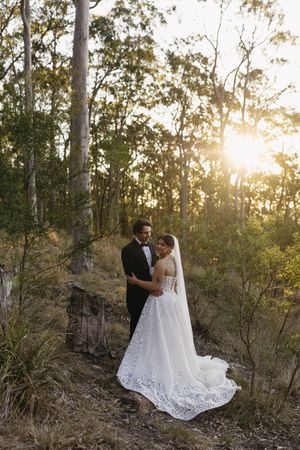 Bride and groom embrace at golden hour in a lush forest, her lace gown trailing elegantly on rocky ground.