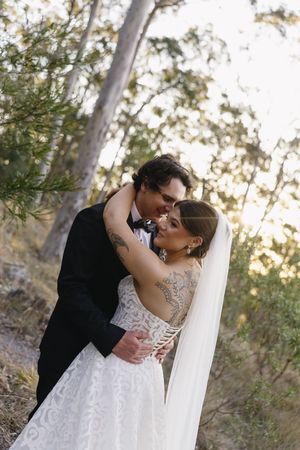 Bride and groom embrace romantically in a forest setting, she wearing a lace backless gown with veil, he in a black tuxedo.