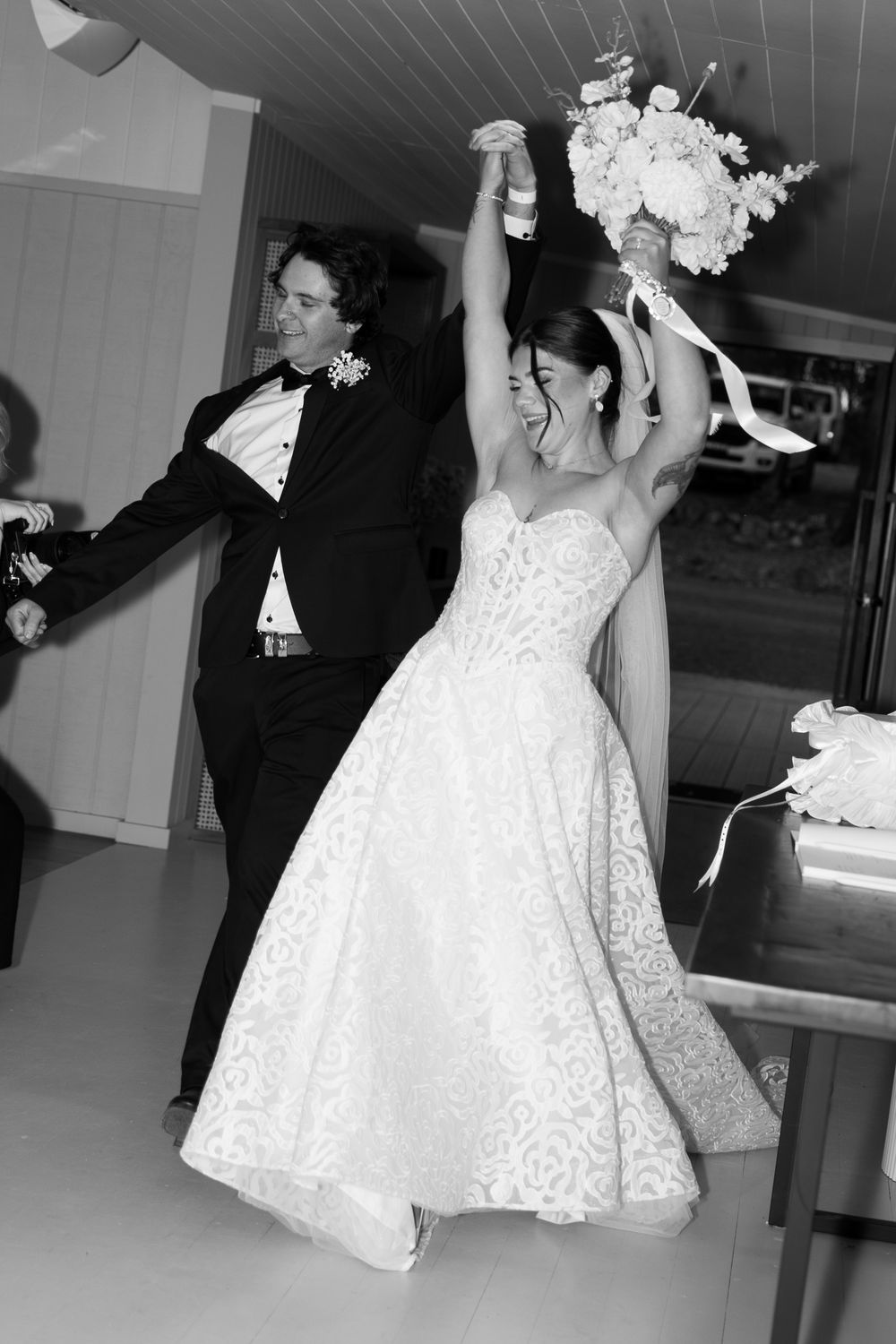 Black and white photo of a joyful bride in lace gown raising bouquet with groom in tuxedo at wedding reception.