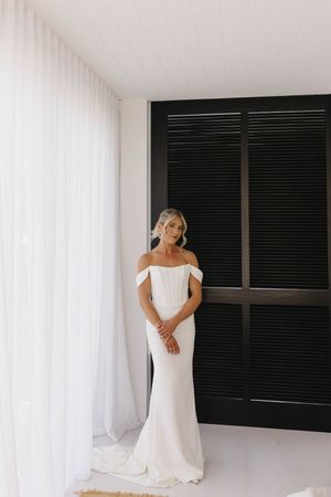 Elegant bride in a fitted off-shoulder white wedding gown poses by a window with modern black shutters.