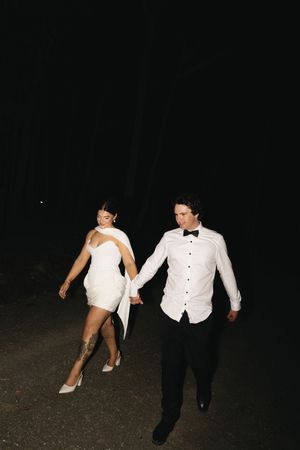 Bride in short white dress and groom in tuxedo shirt holding hands walking at night outdoors.