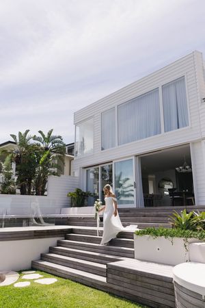Modern white coastal house with wide steps and tropical landscaping against a cloudy sky.