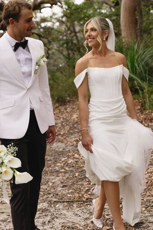 Newlywed couple walking outdoors, groom in white tuxedo jacket and bride in sleek off-shoulder wedding dress.