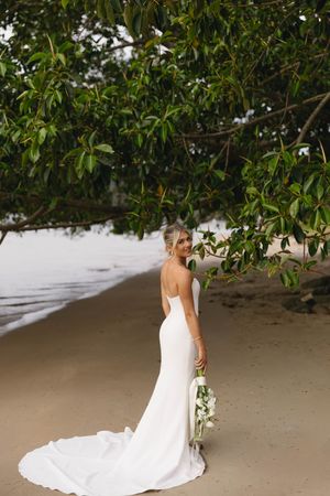 A bride in flowing white dress walks along a sandy beach lined with lush tropical foliage.