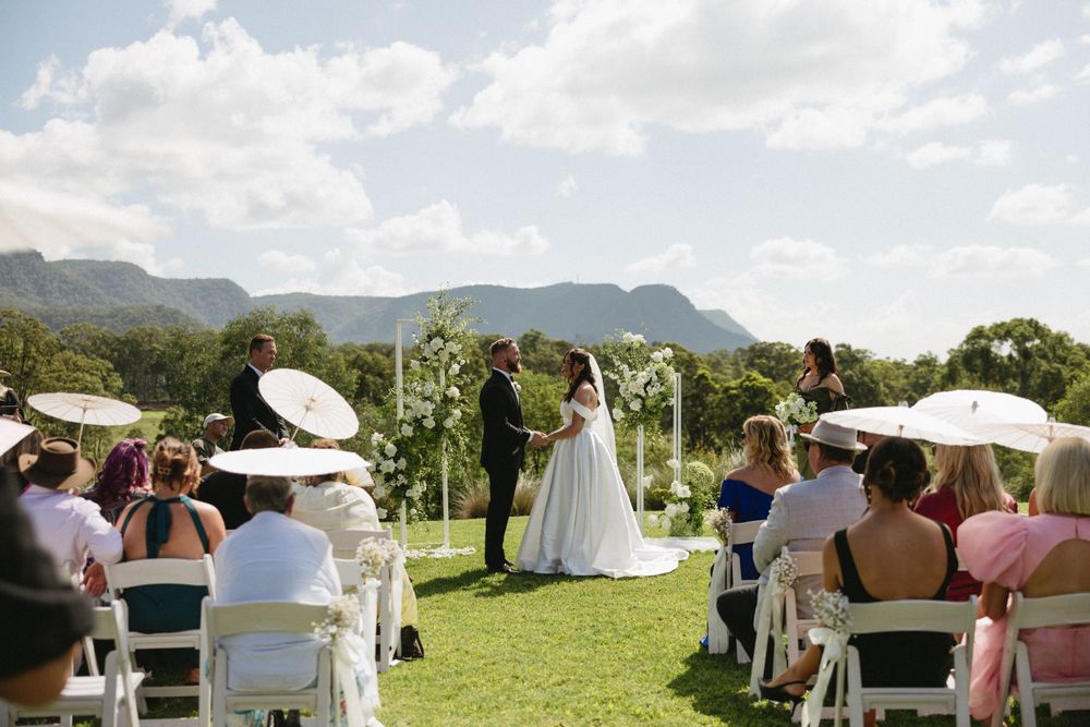 An outdoor wedding ceremony takes place on a lush green lawn with mountain ranges and white umbrellas in the backdrop.