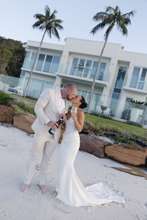 Newlywed couple kissing on white sand beach, groom in cream suit, bride in white gown, holding champagne, tropical palm trees.