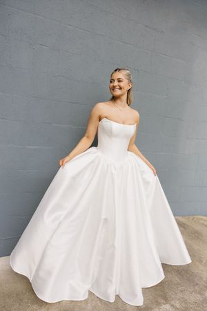 Bride smiling in a strapless white ball gown wedding dress with a full skirt against a gray wall.