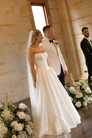Bride in strapless white gown and veil stands with groom in white tuxedo at elegant floral wedding ceremony.