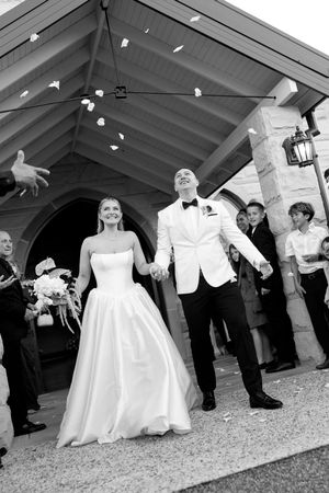 Joyful newlyweds exit church in black and white photo, bride in strapless gown, groom in white tuxedo, guests tossing petals.