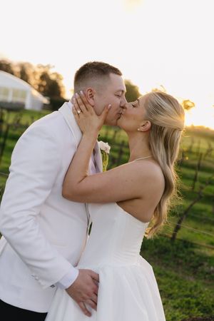 Bride and groom sharing a romantic sunset kiss at a vineyard, wearing white wedding attire.