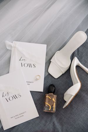 Elegant wedding flat lay with white heels, vow booklets, gold ring, and perfume bottle on gray fabric.