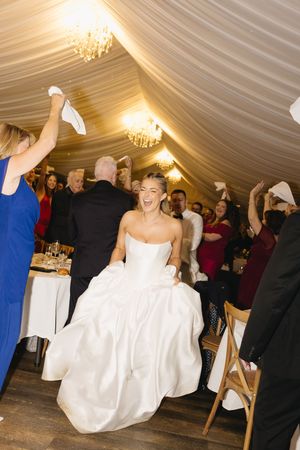 Joyful bride in white strapless gown enters wedding reception tent as guests wave napkins and cheer.
