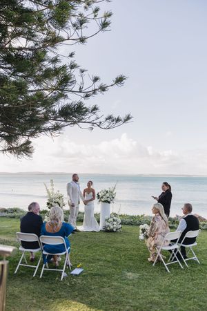 Small intimate outdoor wedding ceremony on green lawn by the ocean with couple at altar, white floral arrangements, and seated guests.