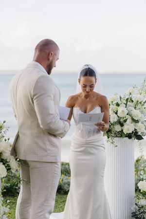 Bride reads wedding vows to groom in white linen suit during outdoor beach ceremony with white floral arrangements.