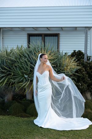 Smiling bride in white mermaid gown and flowing veil poses outdoors near lush green plants by a white building.