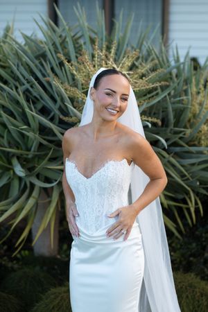 Smiling bride in strapless lace wedding gown and veil poses outdoors in front of lush green plants.
