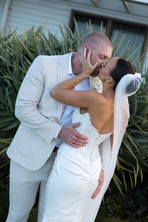 A bride and groom share a romantic kiss outdoors, with the groom in a cream suit and bride in a white lace wedding gown with veil.