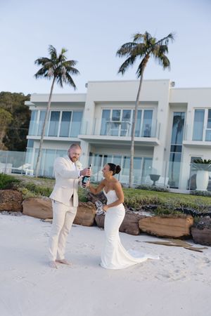 Bride and groom opening champagne on white sand beach beside luxury villa with palm trees.
