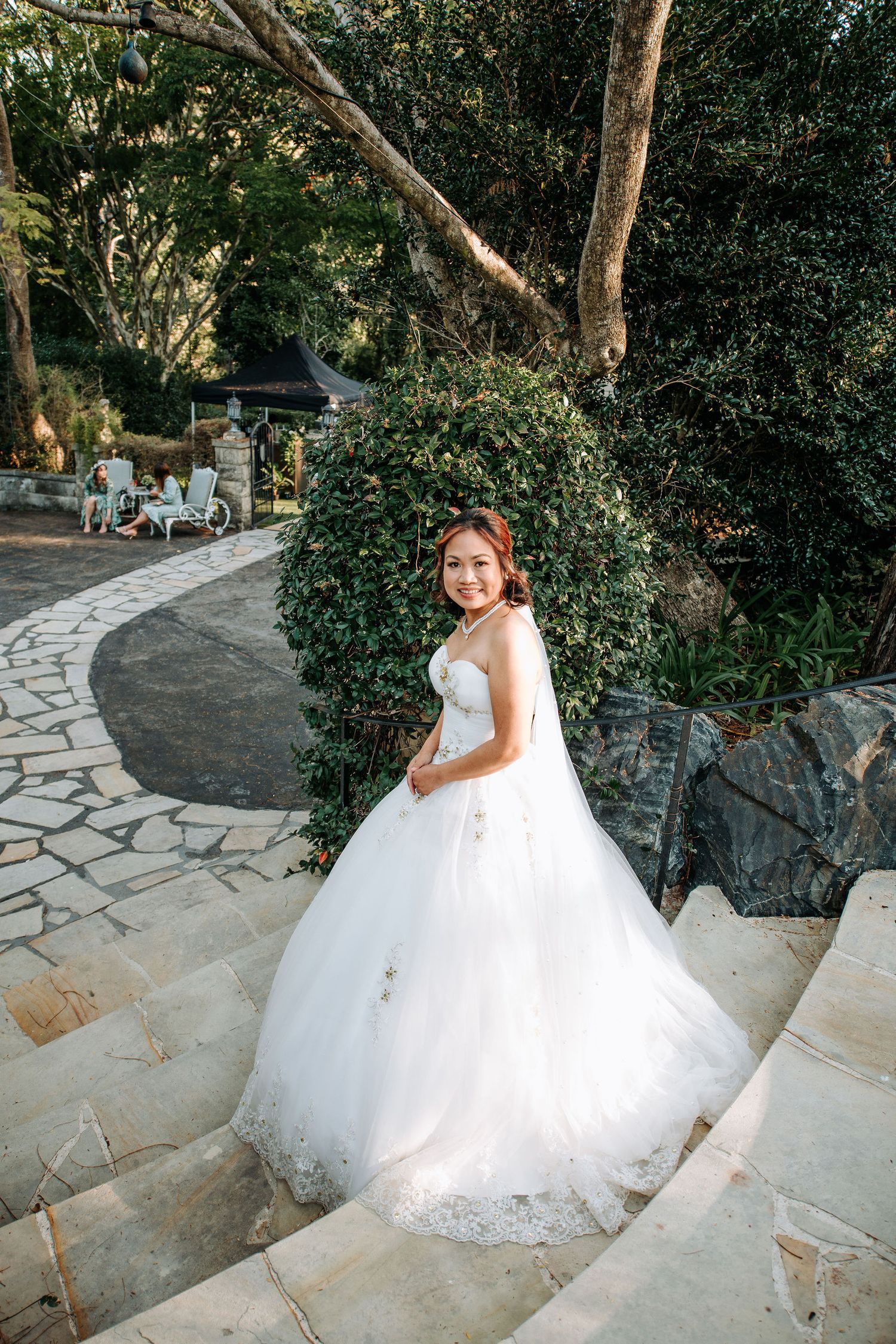 A bride in a princess ballgown poses on stone steps surrounded by lush garden scenery.