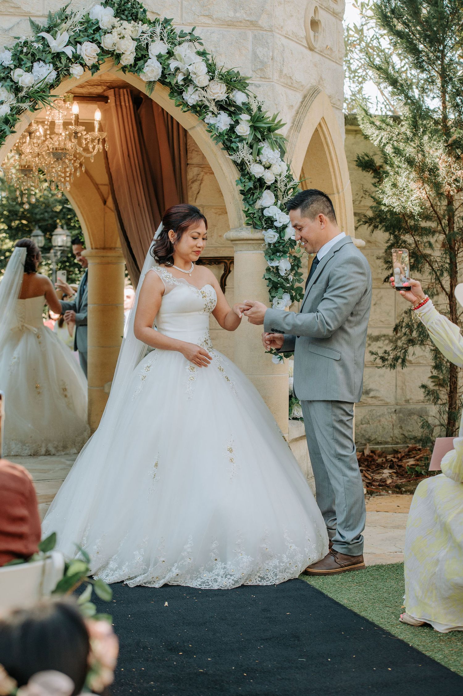 Ring exchange ceremony under an ornate archway adorned with white flowers and greenery.