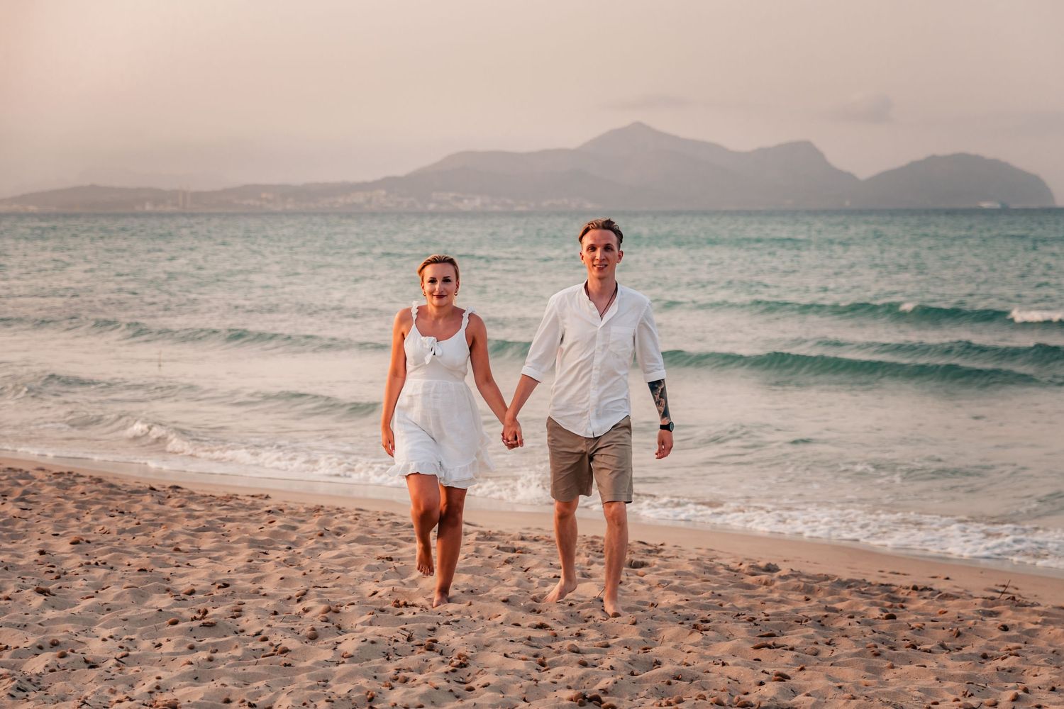 Full length couple walking hand in hand along the shoreline after a surprise beach proposal.