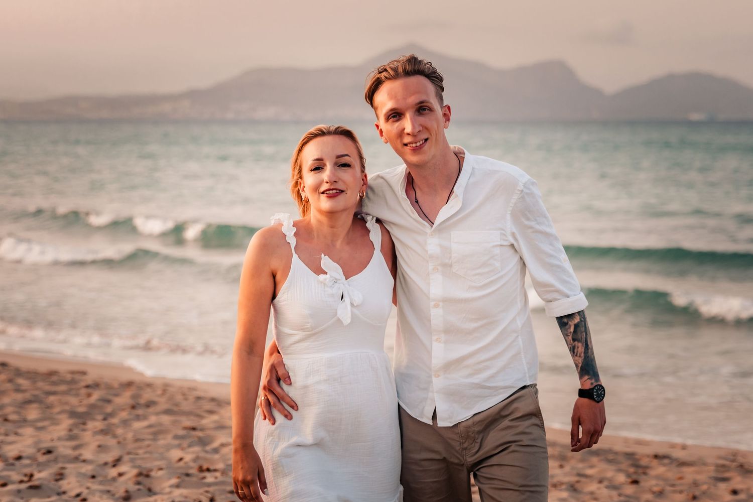 Half length couple walking hand in hand along the shoreline after a surprise beach proposal.