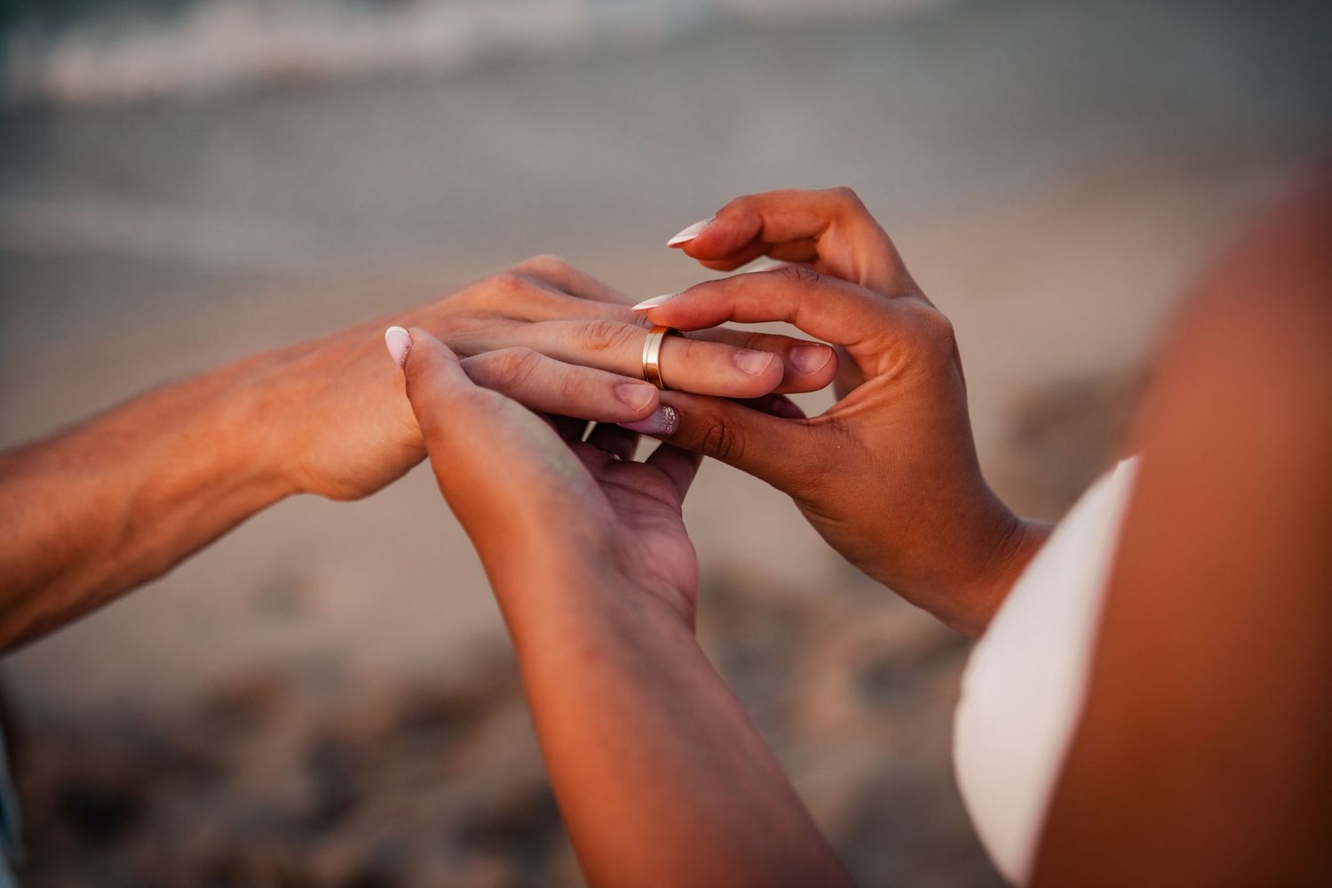 Close-up of a man putting an engagement ring on a woman's finger at a beach by sunset.