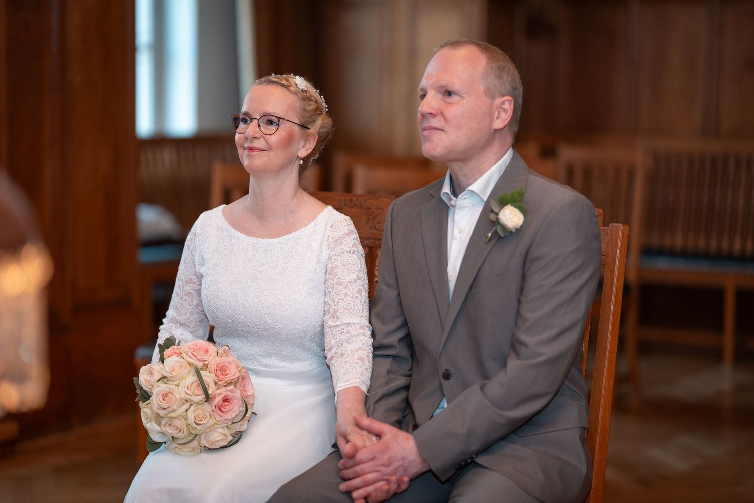 Wedding couple seated together in a formal church venue holding hands while wearing a lace gown and grey suit.