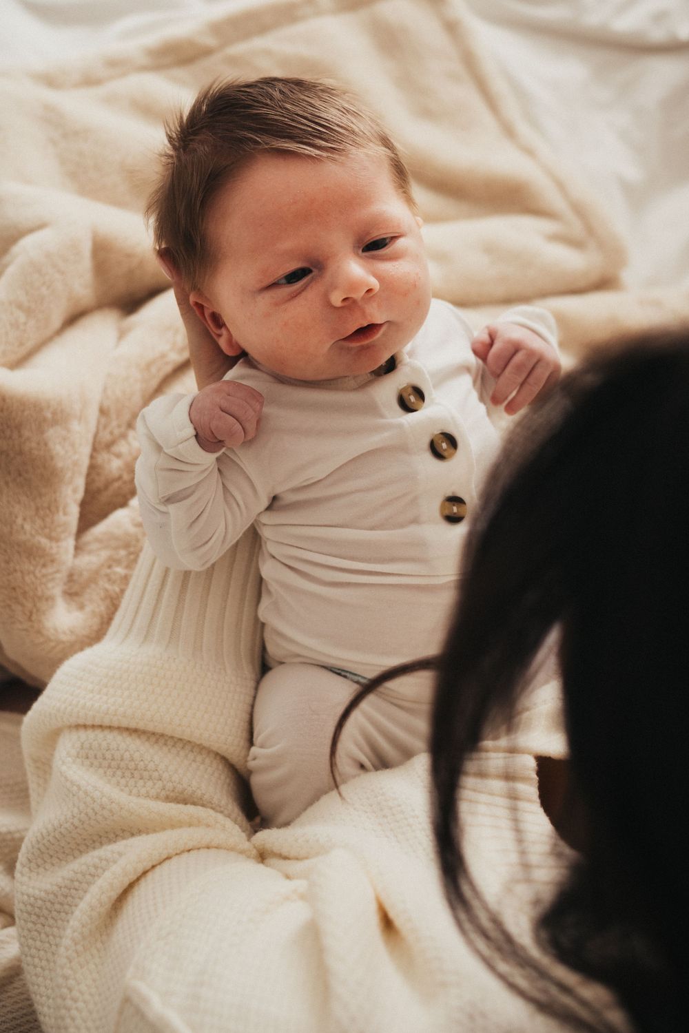 Baby wearing white buttoned romper lying on cream knit blanket looking up at camera.