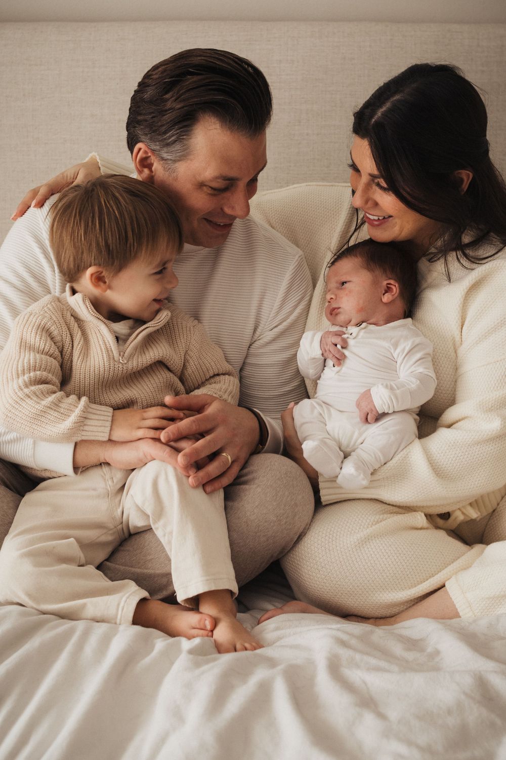 Family of four sharing intimate moment together on bed in coordinated neutral outfits.