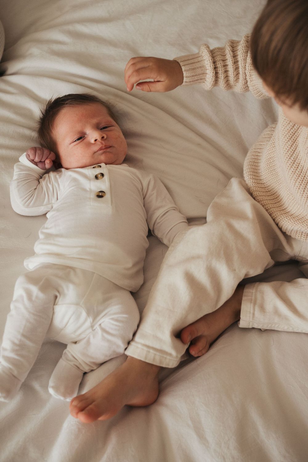 A sleeping infant in white clothing lies on light bedding while being gently touched.