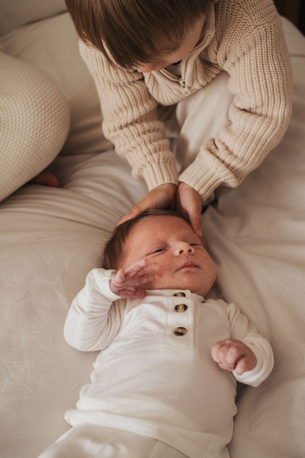 A gentle caress of a sleeping newborn wearing a white buttoned outfit on cream bedding.