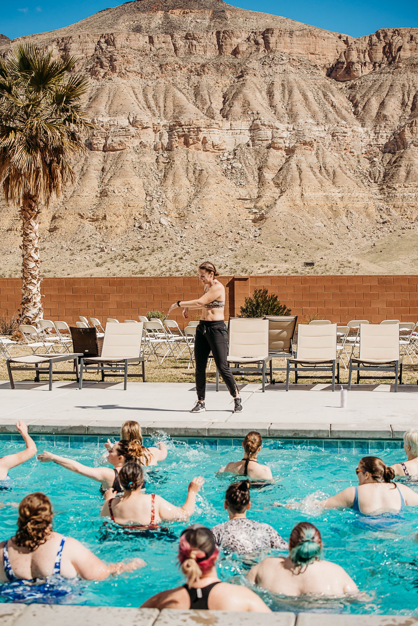 Pool party scene with people in water and person jumping from poolside against desert mountain backdrop.