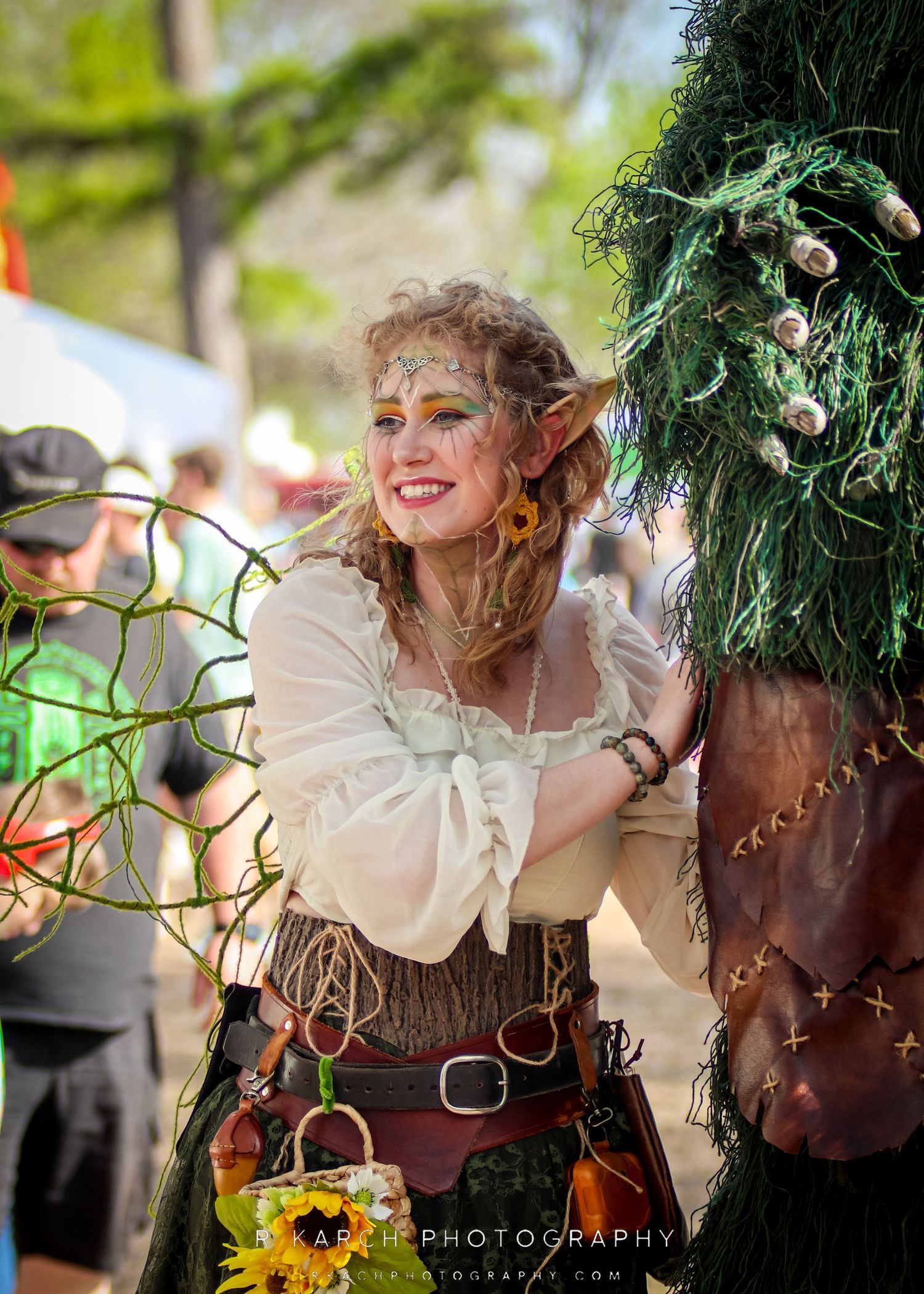 A whimsical performer in renaissance faire attire smiles while posing next to decorative greenery.