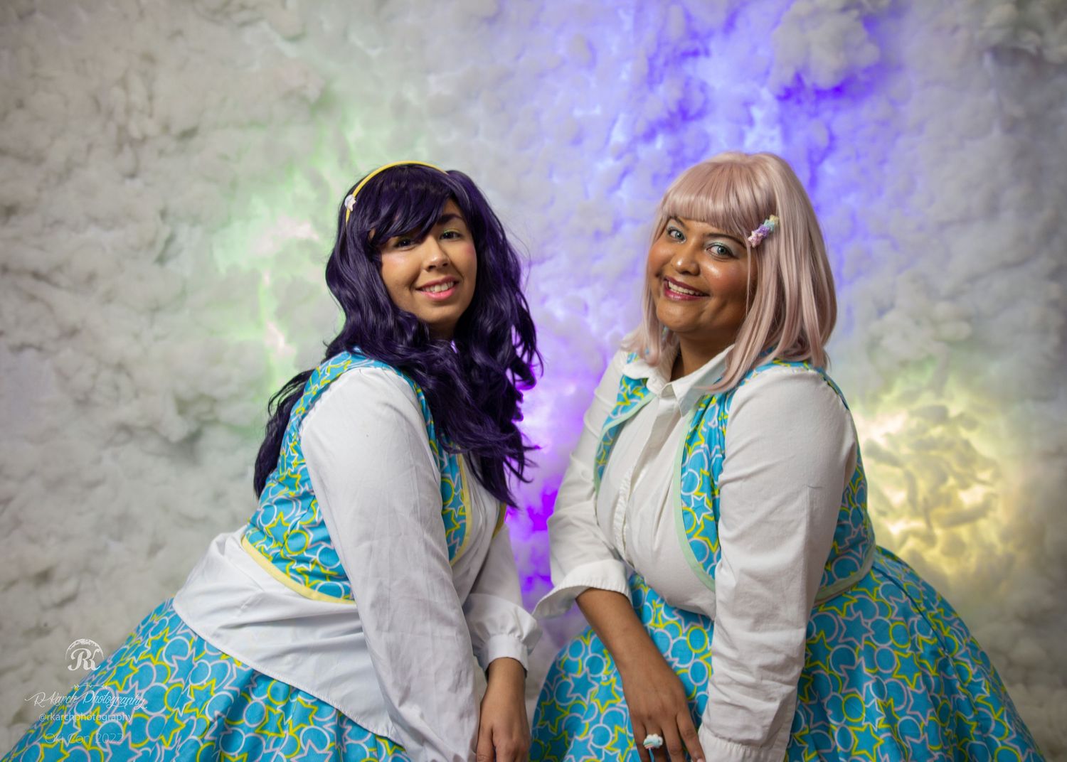 Two cosplayers in matching teal and white patterned dresses pose together.