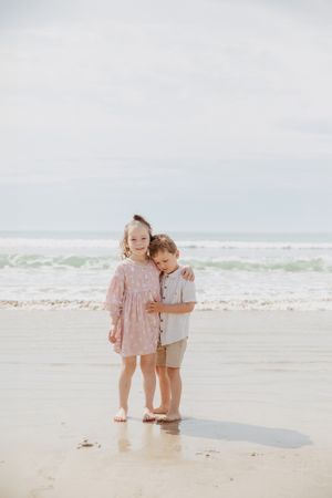 Two children in pastel clothing stand together on a beach with waves in the background.