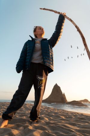 Person playing with a driftwood branch on sandy beach at sunset with seabirds flying overhead.