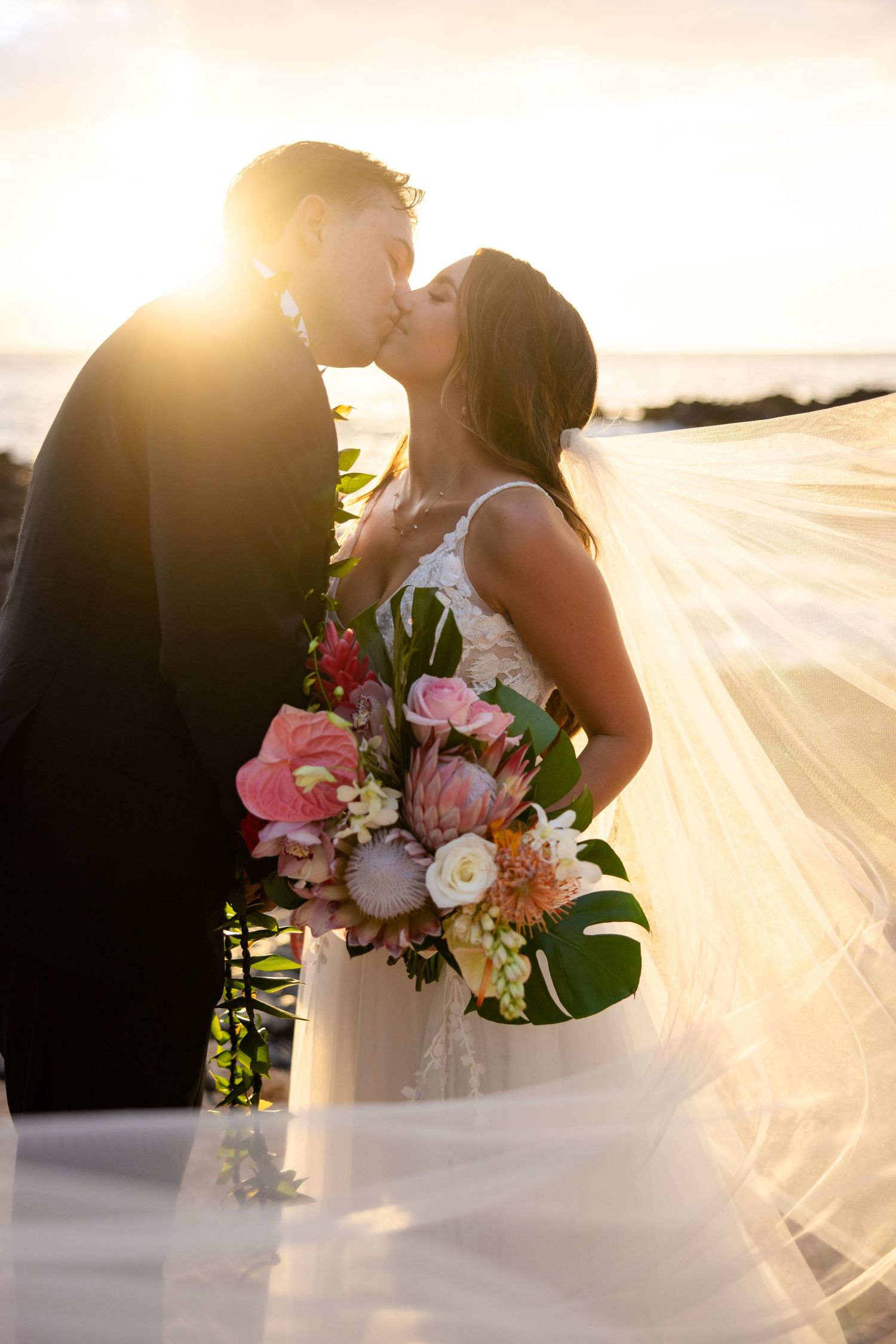 Bride and groom kiss at sunset on the beach, veil flowing, bride holding tropical bouquet.