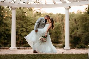 Bride and groom share a romantic dip kiss under a white pergola surrounded by lush greenery at an outdoor wedding.
