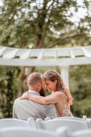 A bride and groom share a romantic forehead touch under a white gazebo surrounded by lush green trees on their wedding day.