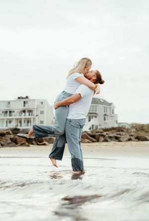 Couple sharing a romantic kiss on a beach, man lifting woman in his arms, with coastal houses in the background.