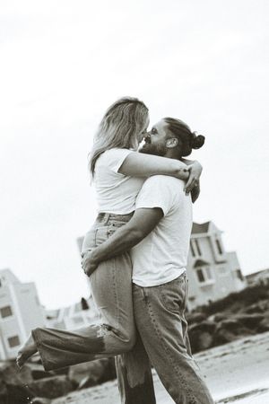 Black and white photo of a couple embracing on a beach, man lifting woman as they lean in for a kiss.