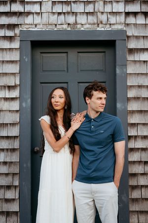 A couple poses in front of a gray shingled house with a dark door, woman in white dress, man in blue polo shirt.