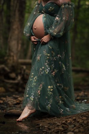 Pregnant woman in floral green tulle gown cradles bare belly outdoors near rocky stream in lush forest setting.